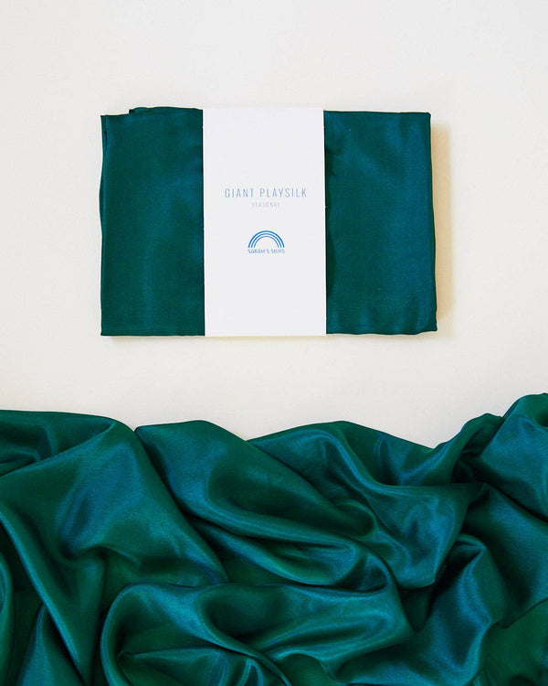 A neatly folded green silk fabric, crafted from luxurious mulberry silk, displays the label "Sarah's Silk Giant Evergreen Playsilk." Below, the same fabric is spread out with gentle folds, highlighting its shiny texture and evoking a hint of winter colorways.