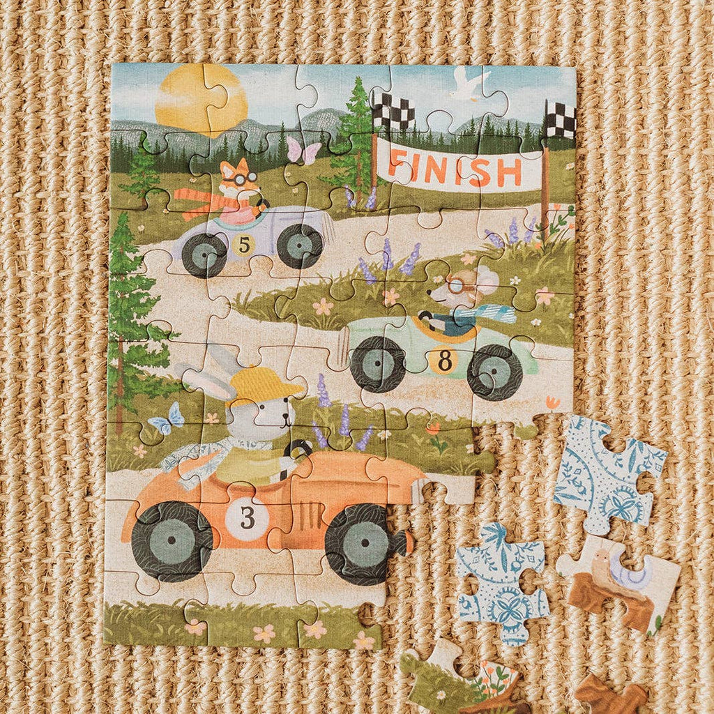 A nearly completed Little Racers 40 Piece Mini children’s jigsaw puzzle on a woven rug shows cartoon animals in cars racing toward a finish line. Three pieces are missing, and some loose pieces from the Little Racers puzzle lie nearby.