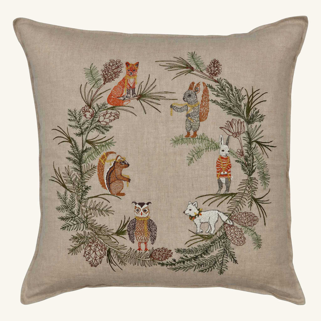 The Coral & Tusk Tree Trimmers Wreath Pillow is a square beige Christmas pillow featuring embroidered woodland animals—fox, squirrel, rabbit, owl, chipmunk, and a white wolf—framed by a pine and cone holiday wreath.