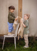 A young girl stands on a chair holding a Senger Naturwelt Cuddly Animal - Linen Goose, facing a boy with a similar toy. They smile at each other while playing outdoors in front of a wooden wall with grass at their feet.