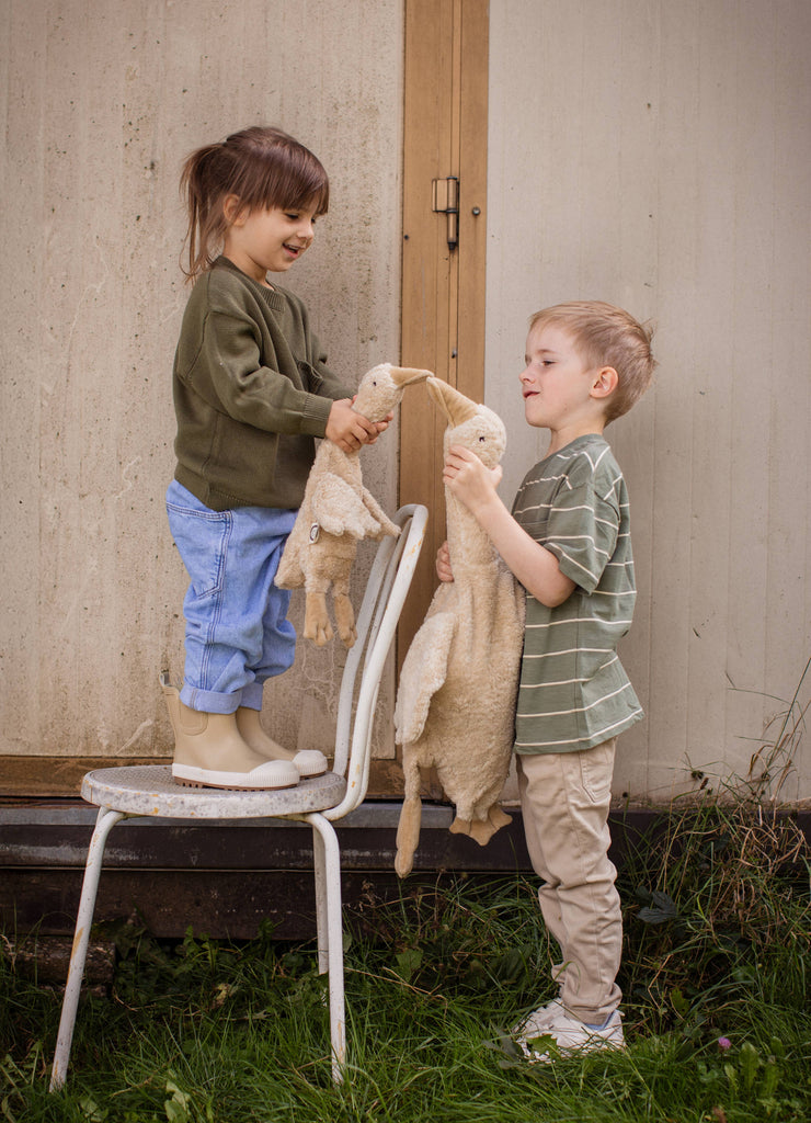A young girl stands on a chair holding a Senger Naturwelt Cuddly Animal - Linen Goose, facing a boy with a similar toy. They smile at each other while playing outdoors in front of a wooden wall with grass at their feet.