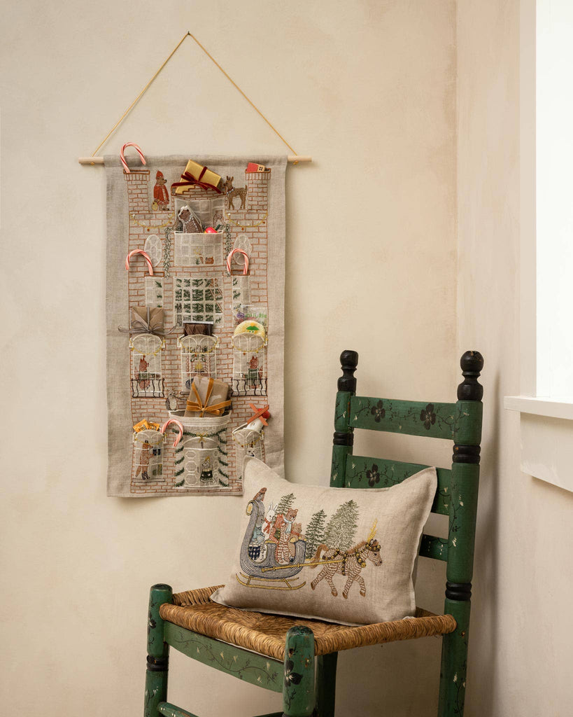 A Coral & Tusk Holiday House Advent Calendar shaped like a brick house with pockets, featuring small gifts and candy canes, next to a chair with an embroidered pillow depicting a horse-drawn sleigh.