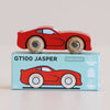 A red wooden sports car rests atop a blue box labeled “No. 18 GT100 Jasper | Made in England.” The box displays an illustration of the car along with icons denoting sustainable wood and child-safe paints, capturing the classic allure of performance motoring in an enduring toy.