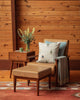 A cozy reading nook with a tan armchair, ottoman, gray throw, Coral & Tusk Downhill Skiers Pillow, wooden side table holding a green mug and vase of flowers, set against wood paneling and an orange rug.