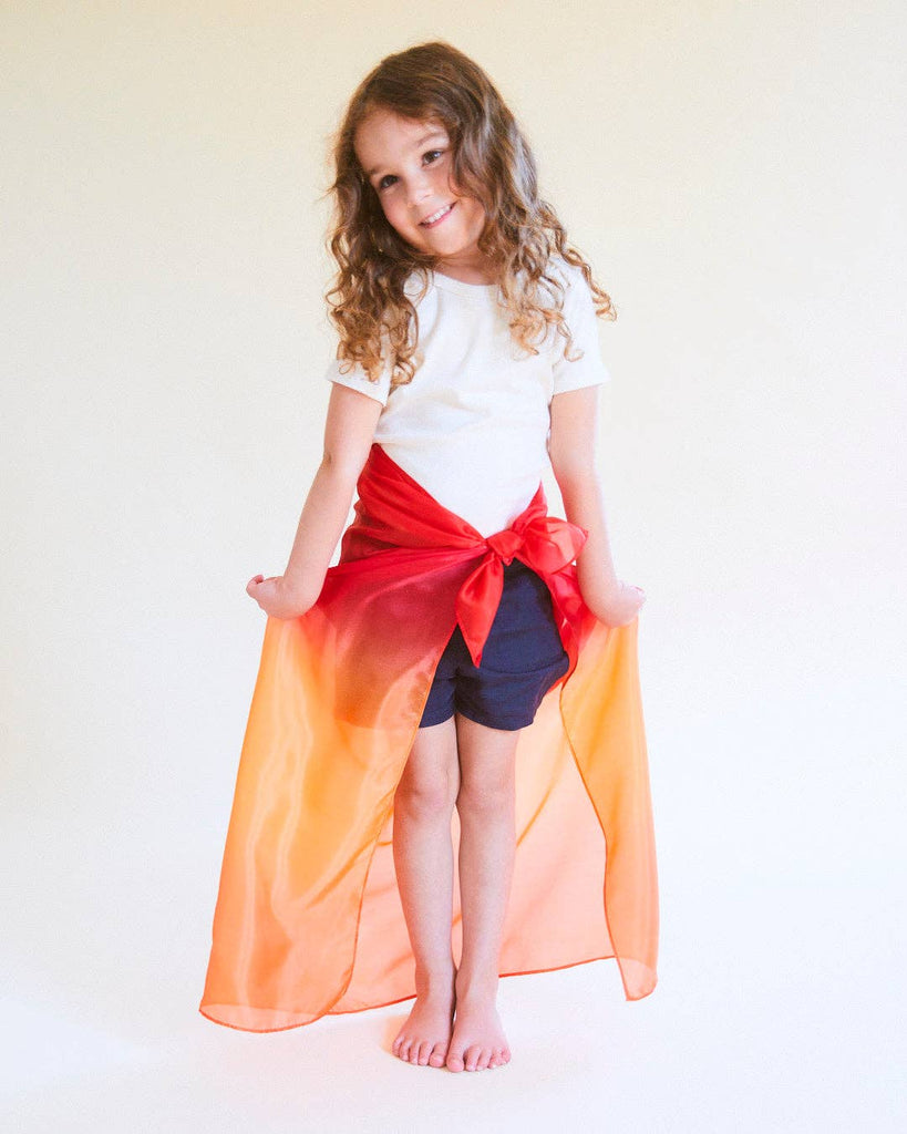 A young child with long curly hair smiles barefoot, wearing a white t-shirt, dark shorts, and Sarah's Silks Enchanted Playsilk - Fire as a vibrant skirt, sparking open-ended play against a plain light background.