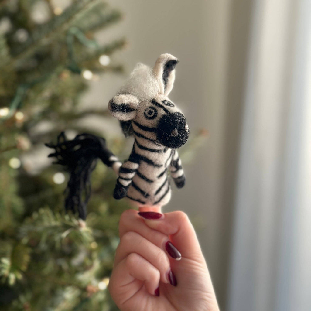 A hand with red nails holds the Zebra Finger Puppet in front of a blurred background featuring a decorated Christmas tree.
