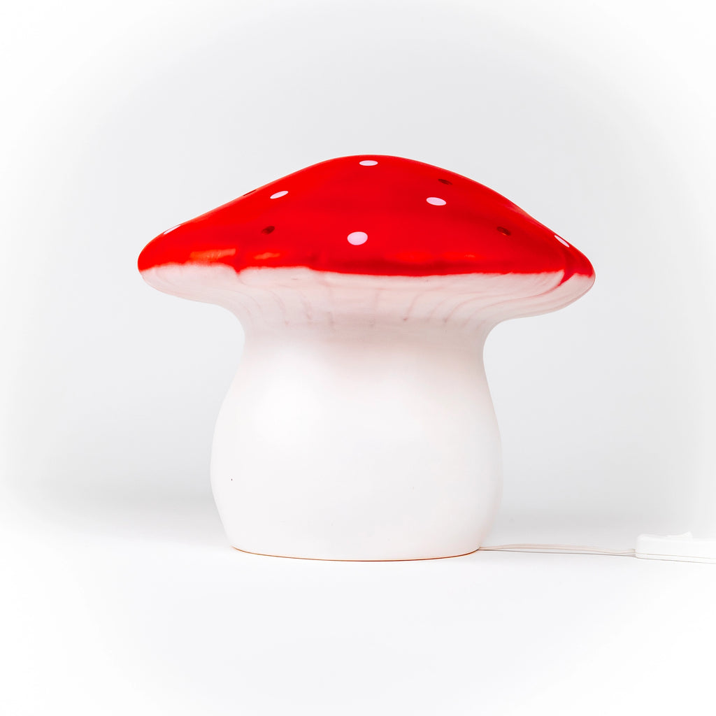 The Red Mushroom Lamp is a whimsical night light with a white stem and red cap featuring small white spots. Plugged in and set against a plain white background, it’s perfect as a nursery lamp or for any children's room.