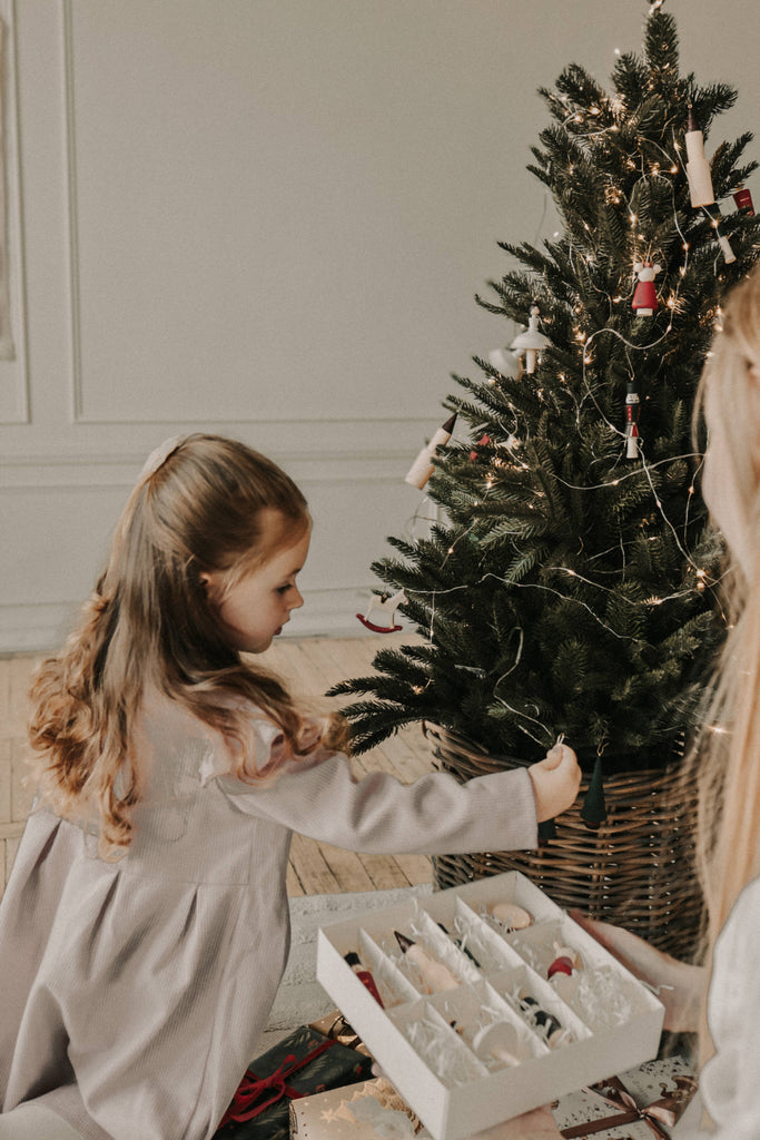 A young girl in pajamas hangs a ballerina ornament on a festive tree as an adult holds a box of Sabo Concept Christmas Ornaments. Wrapped gifts under the tree complete the cozy indoor holiday scene.