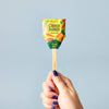 A hand with blue-painted nails holds a Garden Seed Pop (Single) featuring colorful packaging and “Plant Me!” on a wooden stick—great for family-friendly gardening, set against a light blue background.