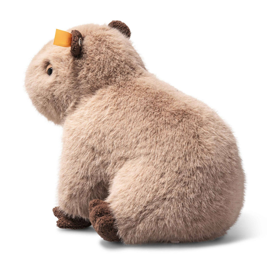 The Steiff Chip Capybara is a plush toy with light brown fur, darker brown feet and ears, and a small yellow rectangle on its left ear, shown sitting on a white background.