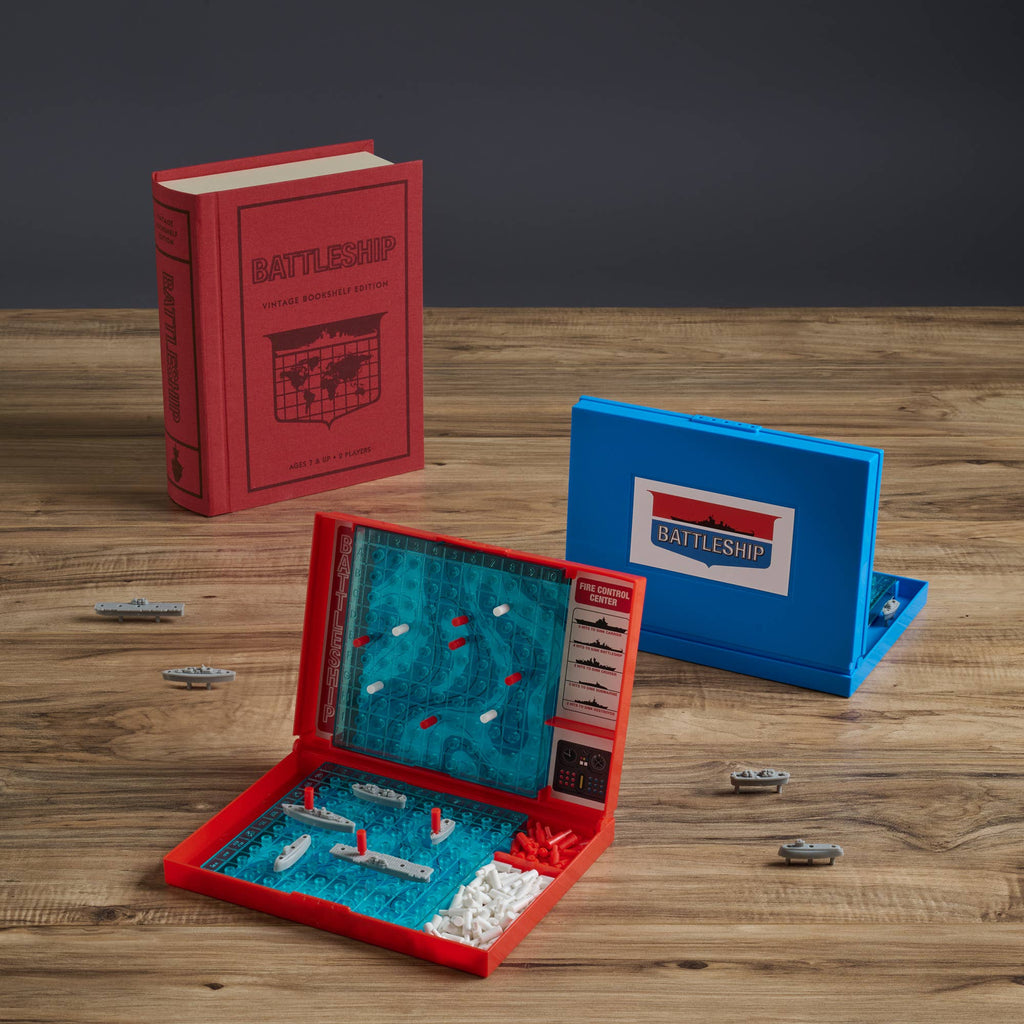 The Board Book: Battleship Vintage Bookshelf Edition is displayed on a wooden table, featuring an open game board with red and white pegs, plastic ships on the grid, a red book-shaped box, and mini ship pieces for classic gameplay.