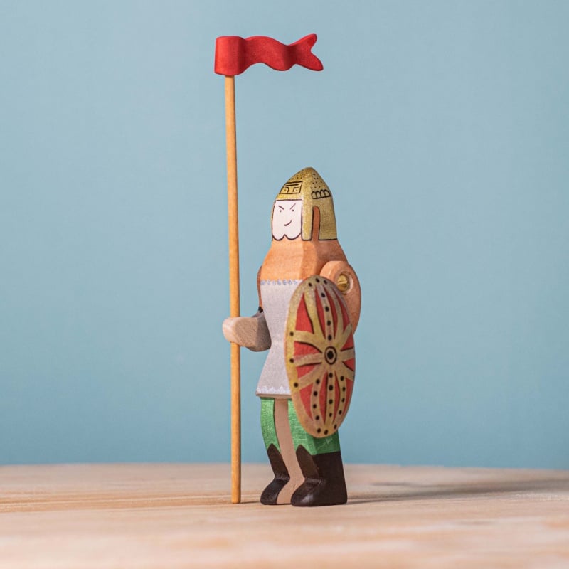 The Bumbu Handmade Wooden Dacian Knight and Steed Set features a handcrafted knight in helmet and chainmail, holding a round shield and tall lance with red flag, displayed on a light wooden surface against a plain blue background.