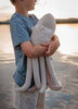At sunset, a child in a blue shirt and light shorts stands by the water, holding a Senger Naturwelt Cuddly Animal - Octopus—a plush organic cotton companion with a removable spelt husk pillow cradled in its arms.