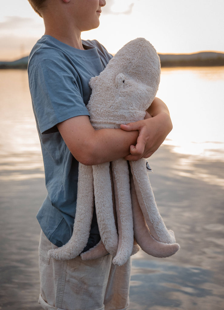 At sunset, a child in a blue shirt and light shorts stands by the water, holding a Senger Naturwelt Cuddly Animal - Octopus—a plush organic cotton companion with a removable spelt husk pillow cradled in its arms.