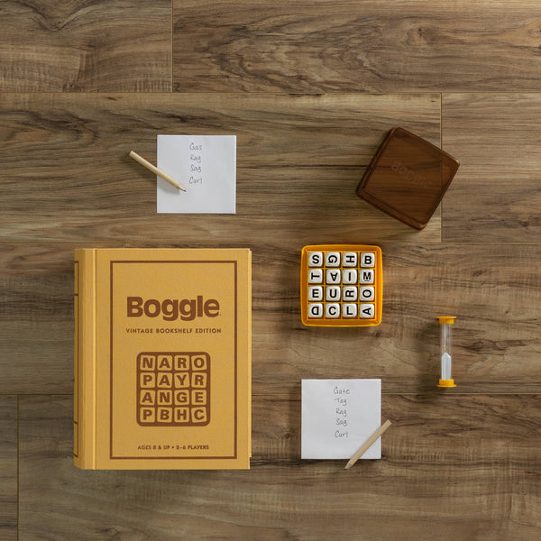 The Board Game: Boggle Vintage Bookshelf Edition is set on wood, showing its box, game cube with letter dice, timer, brown cover, two pencils, and two notepads with handwritten word lists.