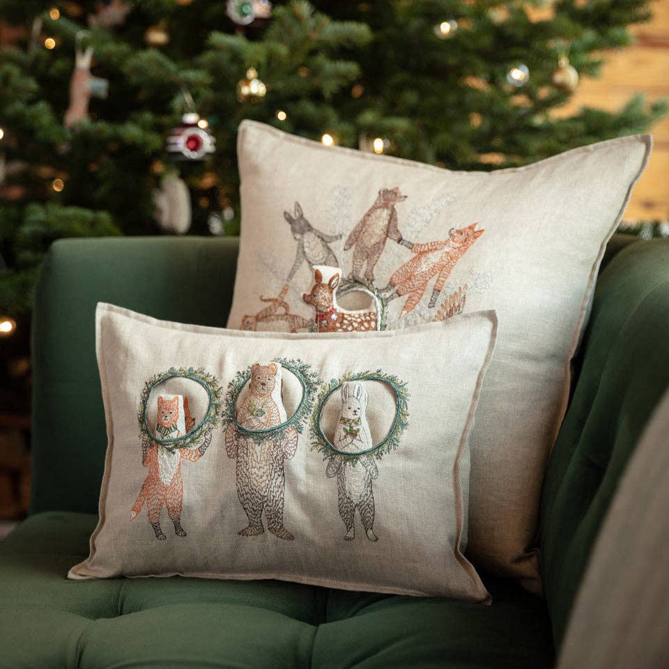 The Christmas Wreath Trio Pocket Pillow, featuring embroidered woodland animals with wreaths, sits on a green couch in front of a Christmas tree, while another pillow behind it shows woodland friends standing together.