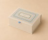 The Delphine Large Embroidered Keepsake Box features a cream-colored rectangular shape adorned with intricate multicolored floral border designs on the lid, accented by a small blue ribbon pull tab on the front, and is displayed against a coordinating beige background.