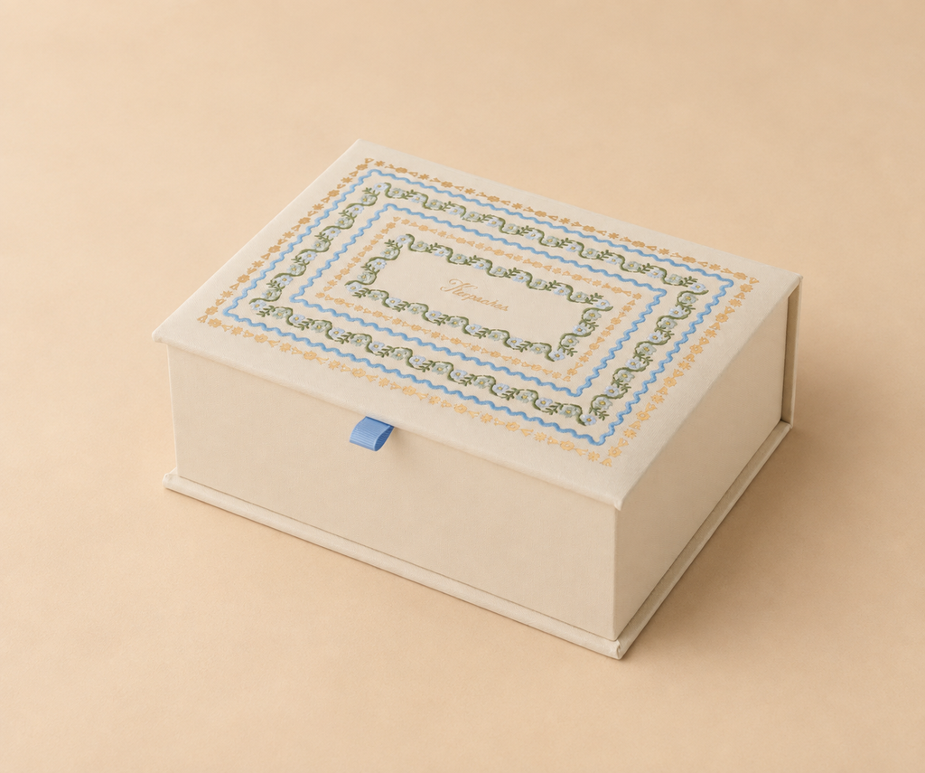 The Delphine Large Embroidered Keepsake Box features a cream-colored rectangular shape adorned with intricate multicolored floral border designs on the lid, accented by a small blue ribbon pull tab on the front, and is displayed against a coordinating beige background.