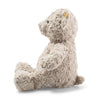 A side view of the Steiff Honey Teddy Bear, 15 inches, sitting. This teddy bear, reminiscent of classic Steiff plush toys, is crafted from light gray, curly textured fabric and boasts a timeless design with a small, rounded tail and ears. It appears soft and cuddly, making it suitable for children or as a collectible item.