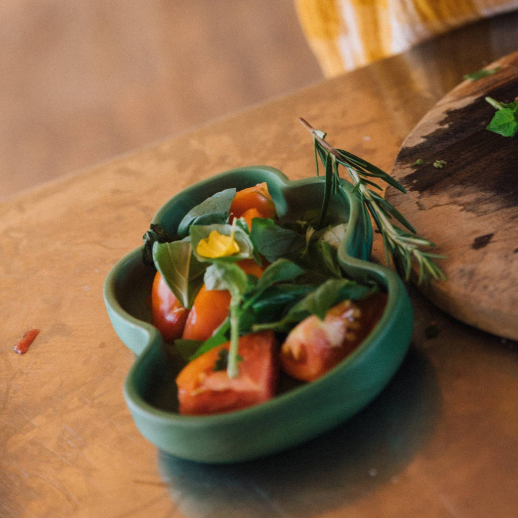 A vibrant green dish from the Kendall the Kale Plate & Spoon Set, presented on a non-toxic plate with tomato slices, fresh greens, and herbs, rests atop a sustainable Hevea wood surface beside a cutting board with scattered leaves—an ideal choice for any baby dinnerware collection.