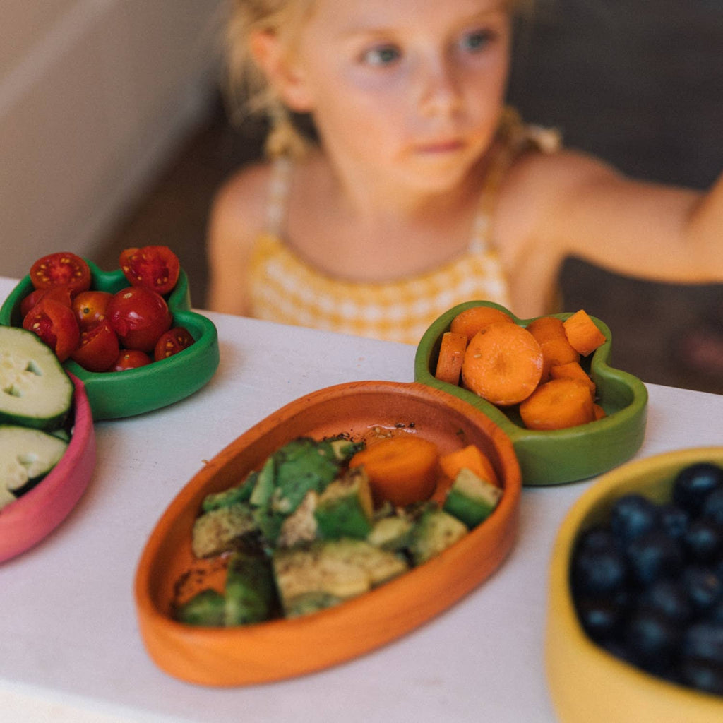 A young child reaches toward colorful bowls filled with fresh vegetables and fruit, including tomatoes, cucumbers, blueberries, and avocado—all beautifully arranged on the Cathy the Carrot Plate & Spoon Set atop a white surface.