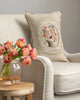 A beige armchair features the Coral & Tusk Sugar Egg Pocket Pillow with embroidered flowers and animals. In front, a small wooden table displays a vase of pink and orange tulips against a softly lit neutral background—perfect Easter decor.