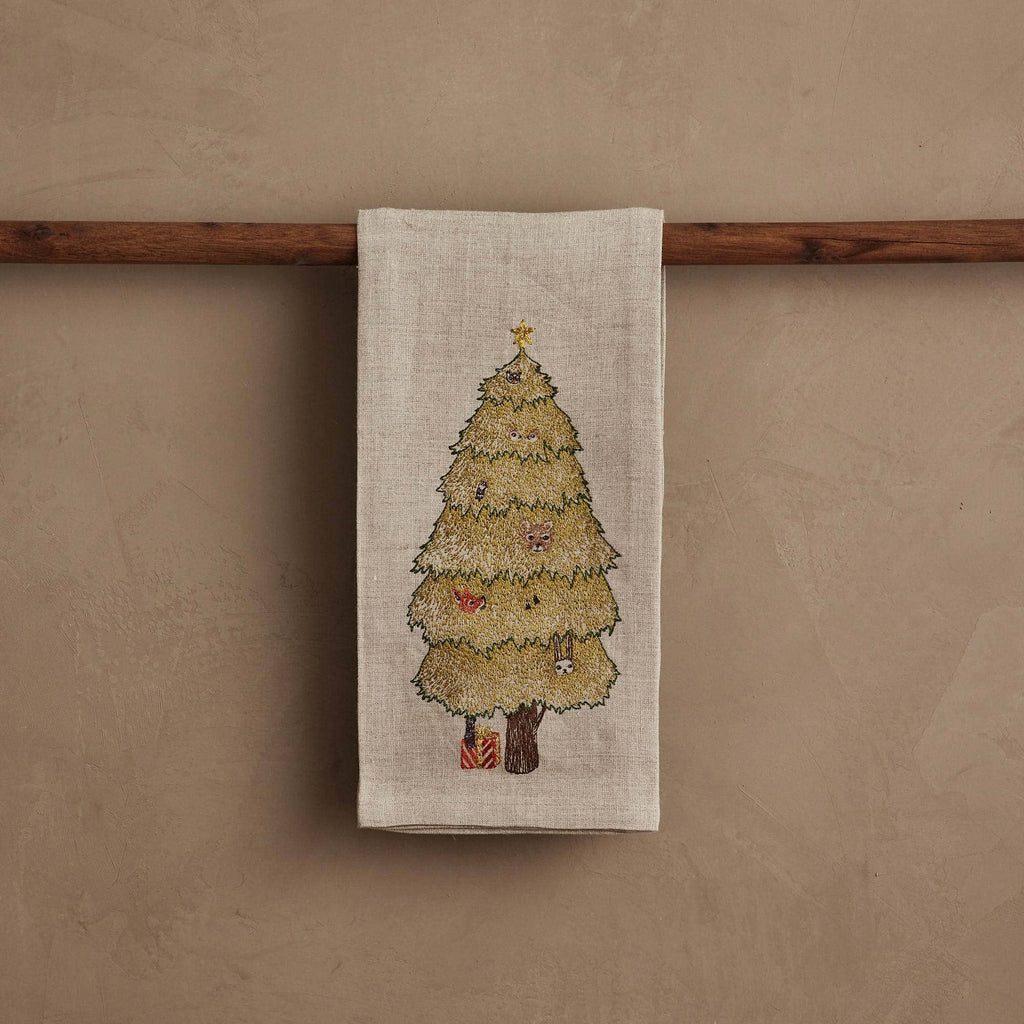 The Coral & Tusk Peek a Tree Tea Towel, in beige linen, hangs on a wooden rod and features an embroidered woodland Christmas tree with ornaments, a shining star, and tiny wrapped gifts—an ideal touch of holiday cheer.