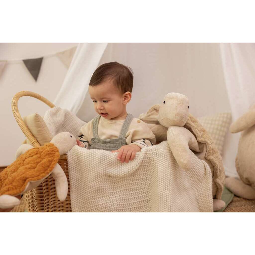 A young child sits in a wicker basket lined with a soft knitted blanket, surrounded by Senger Naturwelt Cuddly Animal - Turtle toys made from organic cotton in a softly lit, neutral-toned room.