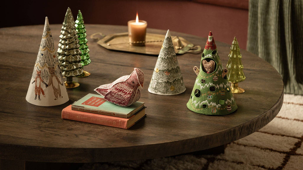 A wooden table displays several Coral & Tusk Peek a Tree Tree felt decorations and a fabric bird, including a green tree with cutouts revealing a bear inside.
