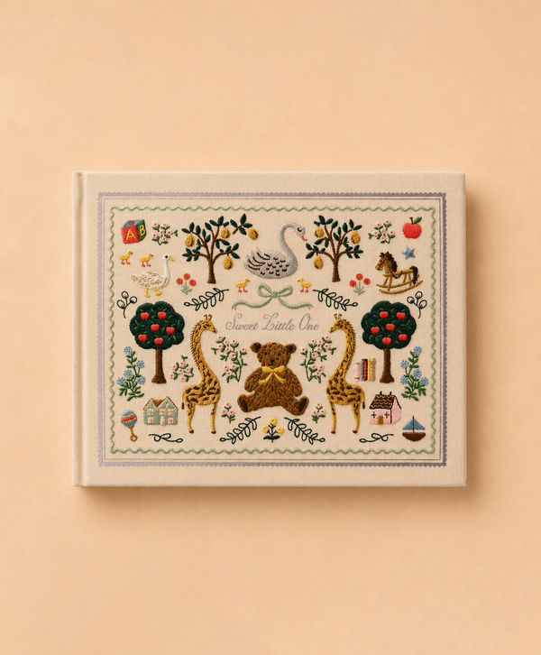 The Storybook Embroidered Baby Book features a cream-colored cover beautifully adorned with embroidered illustrations of a swan, bear, giraffes, trees, animals, and toys. The title “Sweet Little One” is displayed on the pale tan background, making it a charming keepsake for capturing your baby’s precious moments and milestones.
