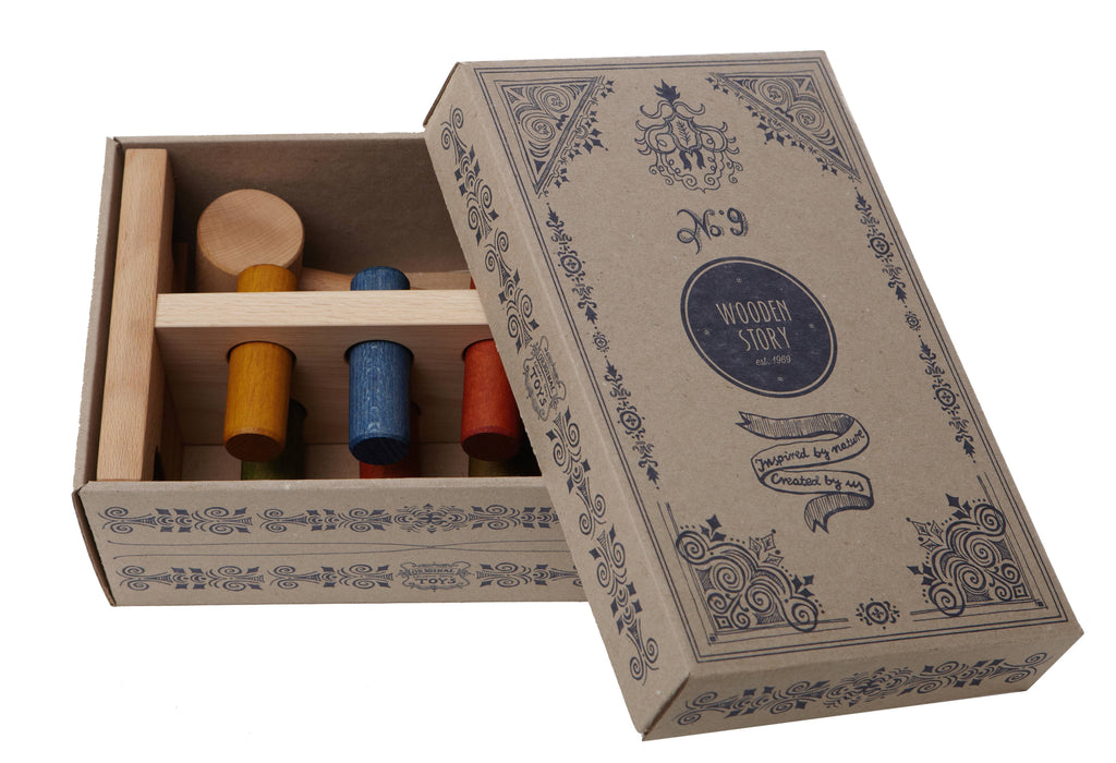 A partially open cardboard box with ornate designs and "No. 9 WOODEN STORY" on the lid reveals brightly colored peg dolls in red, yellow, and blue inside. The Rainbow Pound-a-Peg is a charming eco-friendly toy reminiscent of classic Wooden Montessori toys that inspire imaginative play.