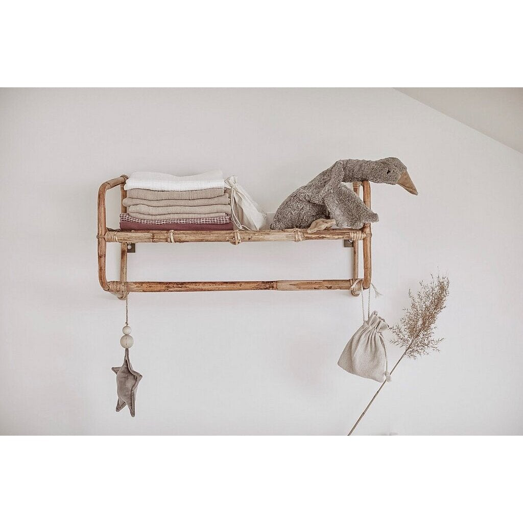 A wooden wall shelf holds neatly folded towels and a Senger Naturwelt Cuddly Animal - Grey Goose. Below, a star ornament, drawstring pouch, and dried plant hang from the shelf against a white wall.