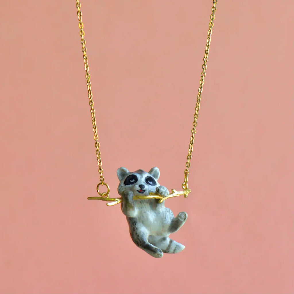 A small gold necklace features a handcrafted pendant of a raccoon figurine hanging onto a small branch, set against a peach-colored background. The raccoon, with its detailed black and white facial markings, clings to the branch, creating a playful appearance. This charming Rascal Raccoon Necklace is attached to an elegant gold chain.