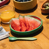The Wally the Watermelon Plate & Spoon Set, featuring a green Hevea wood plate with sliced watermelon, rests on a wooden table beside a green spoon, non-toxic tableware, orange juice, and other colorful dishes.