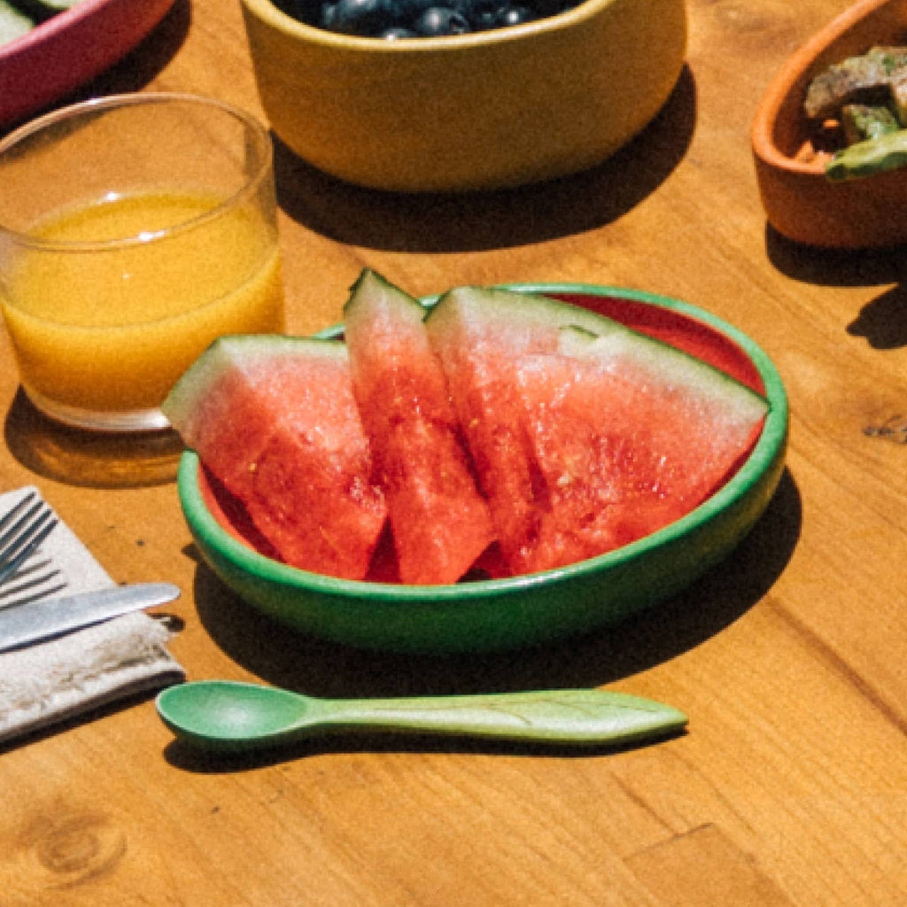 The Wally the Watermelon Plate & Spoon Set, featuring a green Hevea wood plate with sliced watermelon, rests on a wooden table beside a green spoon, non-toxic tableware, orange juice, and other colorful dishes.