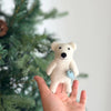 A hand holds the Finger Puppet: Polar Bear Catching a Fish ornament, featuring a white polar bear with a blue ball near its paw, in front of a decorated Christmas tree—ideal for imaginative play and Arctic storytelling.