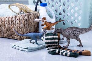 The Schleich Carcharodontosaurus Dinosaur Toy, featuring a movable jaw, is displayed alongside three other dinosaur figures amid laundry room essentials like folded towels, striped socks, a laundry basket, and a detergent bottle to create a playful scene on the floor.