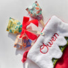 A Pollinator Seed Pop (Single) with a vibrant floral design and planting instructions is displayed by a red bow and a Christmas stocking embroidered with "Owen." Perfect for gifting, it makes growing a pollinator garden simple and fun.