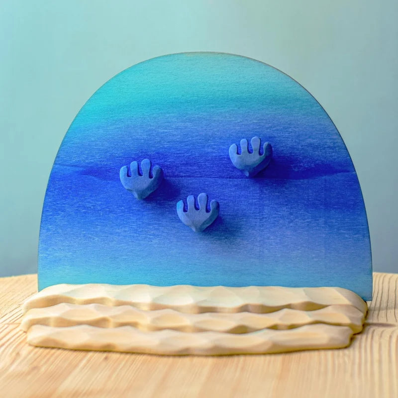 The Bumbu Handmade Wooden Deep Ocean Water Set includes three blue hand-shaped forms rising from wave-like bases, a gradient blue semicircle background, and wavy beige sand pieces, perfect for imaginative play and display.