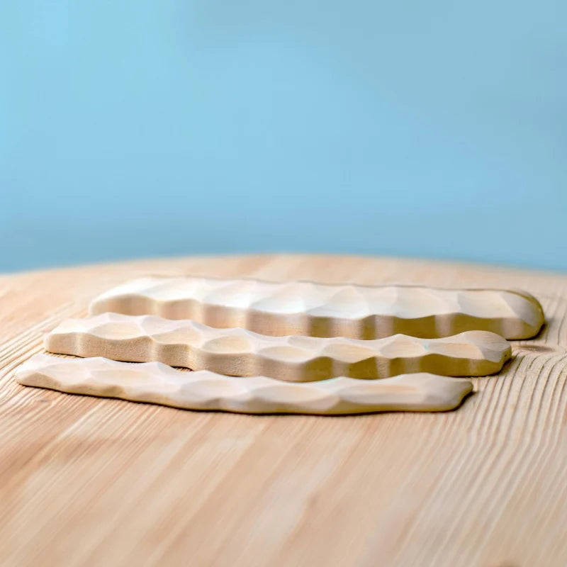 Three pieces from the Bumbu Handmade Wooden Deep Ocean Water Set, with wavy surfaces, rest on light wood against a soft blue background—ideal for imaginative play or stylish display.