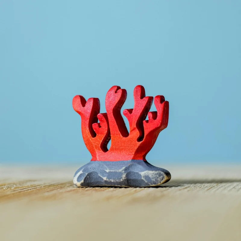 A small red coral figurine from the Bumbu Handmade Wooden Deep Ocean Water Set stands on a painted gray base against a light blue background and wooden surface, crafted from sustainable wood.