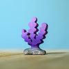 A small, purple coral figurine from the Bumbu Handmade Wooden Deep Ocean Water Set stands upright on a stone base, crafted from sustainable wood, set against a light blue background and wooden surface.