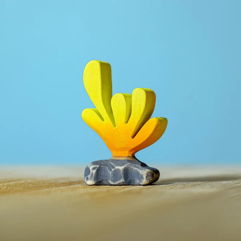 A small yellow and orange coral figurine from the Bumbu Handmade Wooden Deep Ocean Water Set, crafted from sustainable wood with a gray base, stands on a light wooden surface against a blue background.