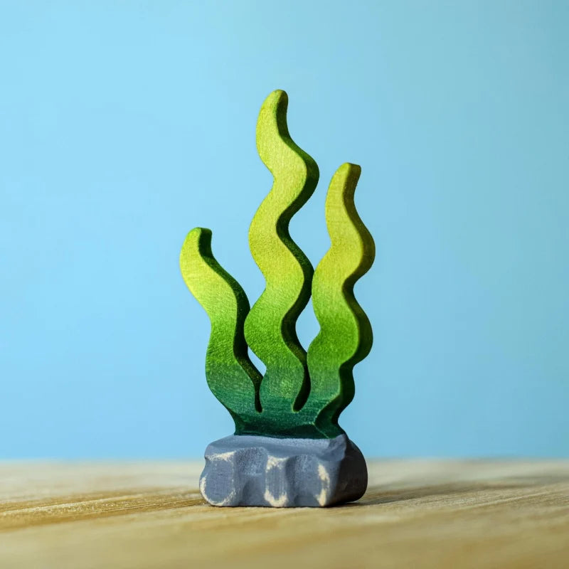 A small, hand-painted wooden seaweed figurine with green fronds stands on a gray rock base—a delightful addition to your Bumbu Handmade Wooden Deep Ocean Water Set for imaginative marine play.