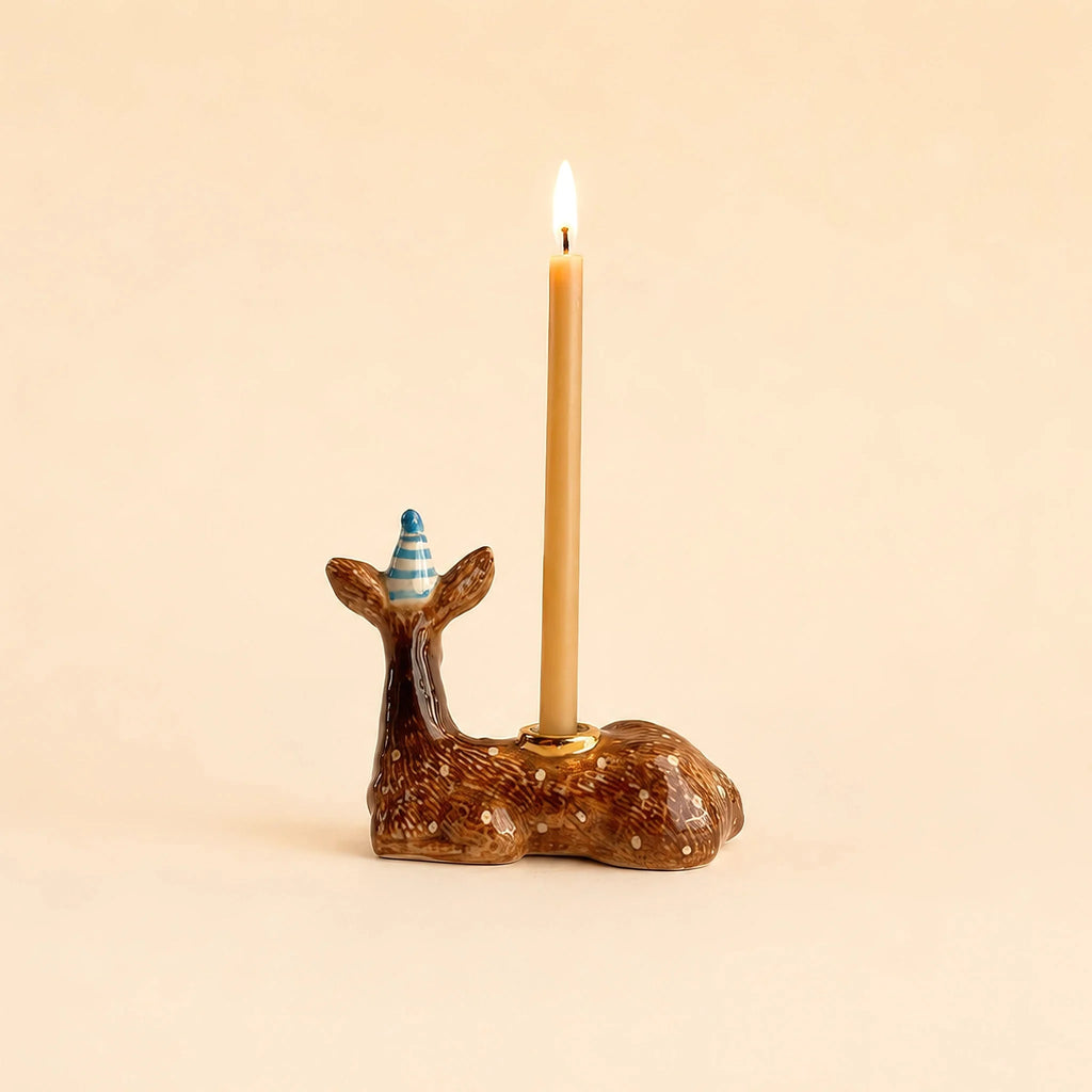 The Deer Cake Topper - Final Sale (ships in one week) is a handcrafted brown ceramic deer with gold accents, wearing a blue party hat and holding a lit taper candle on its back.