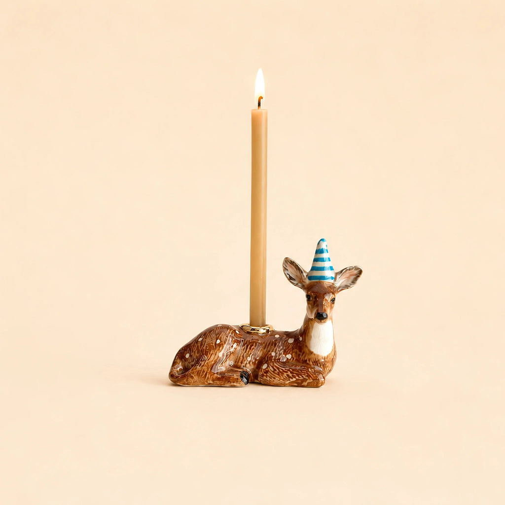 The Deer Cake Topper - Final Sale (ships in one week) is a brown ceramic deer with gold accents and a blue striped party hat, holding a lit beige candle on its back against a plain beige background.