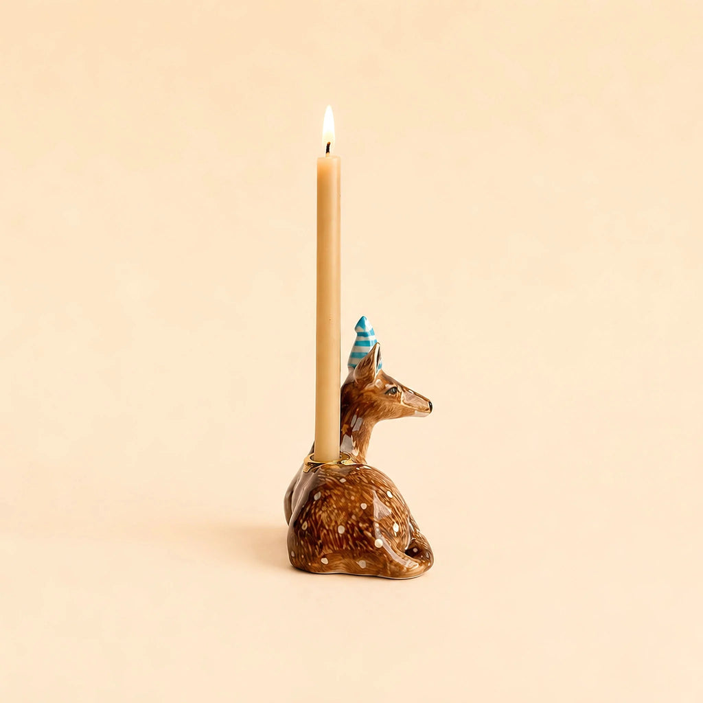 A lit candle sits in the Deer Cake Topper - Final Sale (ships in one week), a brown deer with a blue striped party hat and gold accents, against a beige background.