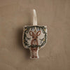 The Coral & Tusk Togetherness Deer Ornament is a fabric ornament featuring a fawn within a wreath of pine branches and baubles, finished with a cream-colored hanging loop and shown on a textured beige background.