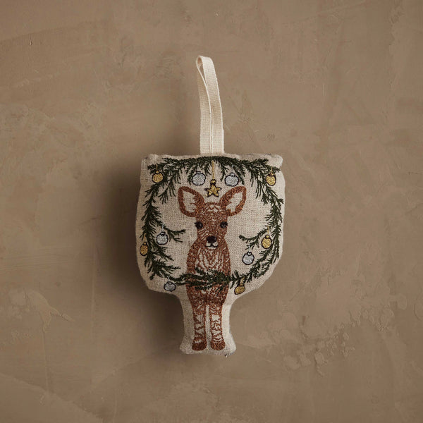 The Coral & Tusk Togetherness Deer Ornament is a fabric ornament featuring a fawn within a wreath of pine branches and baubles, finished with a cream-colored hanging loop and shown on a textured beige background.