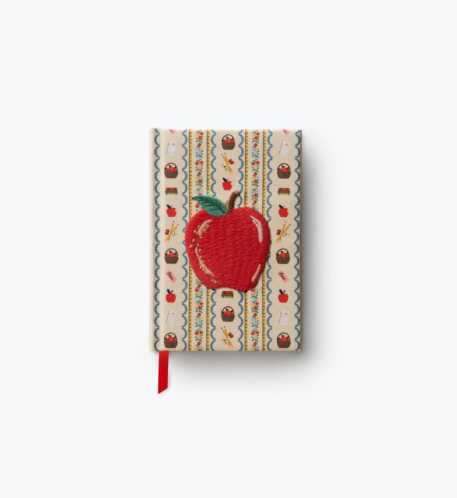The Schoolhouse Mini Embroidered Journal is a hardbound notebook adorned with a large embroidered red apple on the cover and embellished with a colorful, decorative border featuring small icons and patterns. This charming mini journal includes unlined pages for creative expression, along with an attached red ribbon bookmark at the bottom.